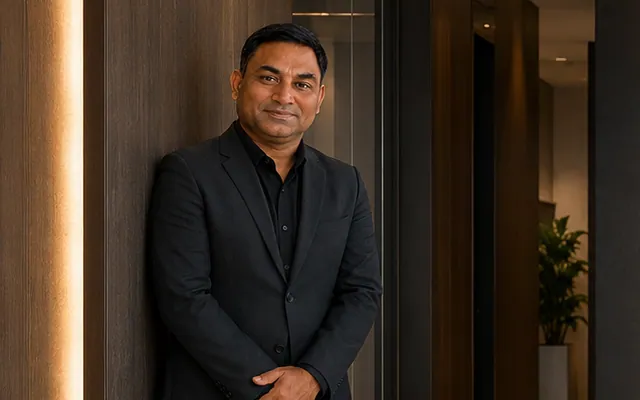 Rohit in a dark business suit leaning against a wood-panelled corridor wall